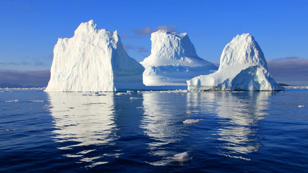 Three bright icebergs reflected in calm blue water beneath a clear sky.